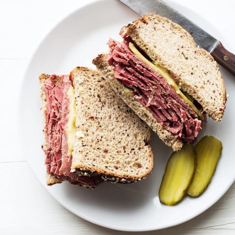 Thick cut of Pastrami on Rye sandwich served on a plate with a crunchy dill pickle spear on the side.
