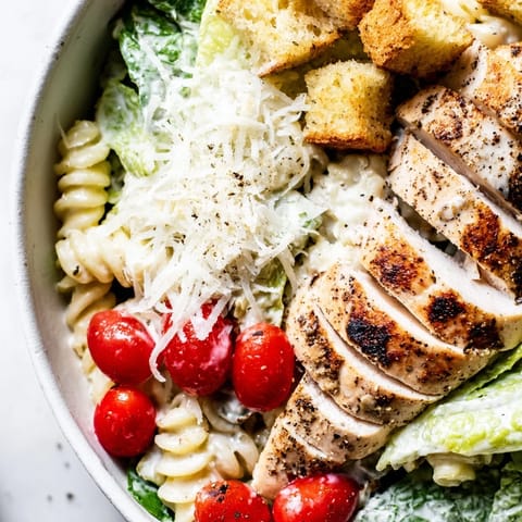 Freshly grilled chicken slices rest atop a vibrant Caesar pasta chicken bowl, mingling with crisp romaine and garlic croutons.