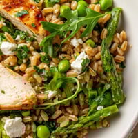 A close-up of Warm Spring Farro Chicken Salad with grilled chicken slices, blanched asparagus, peas, and feta on a bed of farro and arugula.